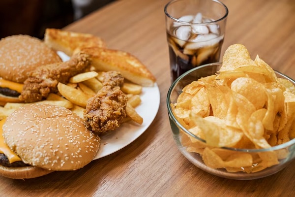 cómo saber si sufro de alopecia - mesa de madera con un plato con dos hamburguesas, dos muslitos de pollo rebozados y patatas fritas, más un bol de patatas chip y un vaso con un refresco de cola y hielo