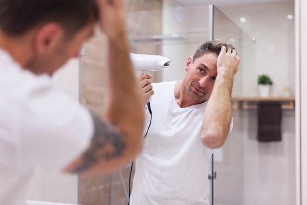 Cómo saber si sufro de alopecia - hombre adulto con una camiseta blanca secando su cabello con un secador delante del espejo del baño