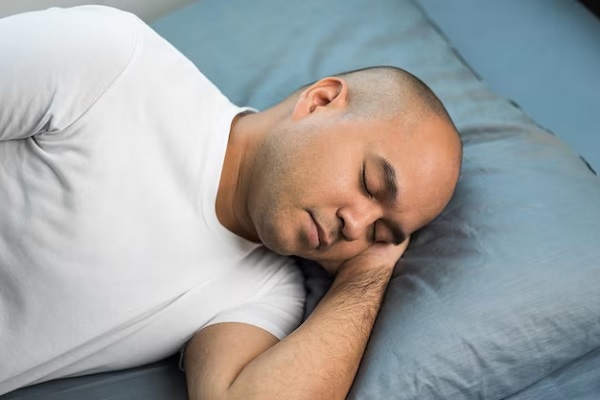 imagen de un hombre en la cama durmiendo de costado con la mano en la cara para que la cabeza no toque la almohada