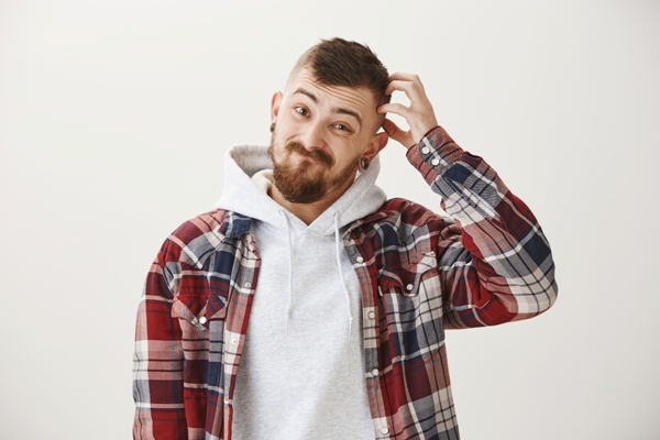 Tipos de entradas en el cabello - Joven de camisa de cuadros se rasca la cabeza en la zona de la sien al notar los primeros síntomas de alopecia