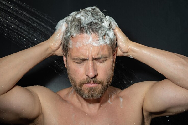 Nutrición para el pelo - Hombre barbado masajea su cuero cabelludo bajo la ducha mientras distribuye el champú sobre su pelo 