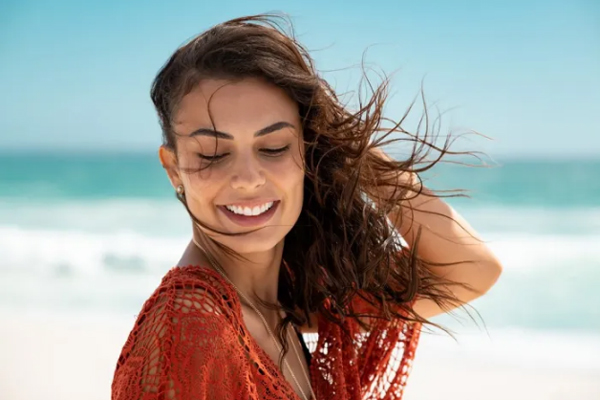 Revitalizar el pelo después del verano - Mujer sostiene su cabello castaño para evitar que se le tape por completo el rostro por la brisa del mar