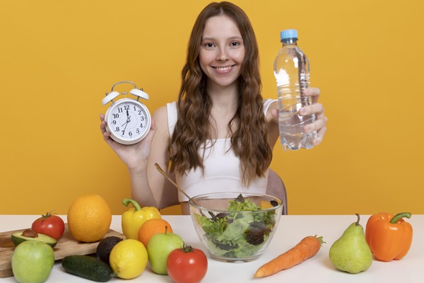 Mujer sentada con una mesa llena de frutas, sosteniendo una botella de agua y un reloj, como mensaje de que es tiempo de comer sanamente, para mantener un cabello y cuerpo saludable