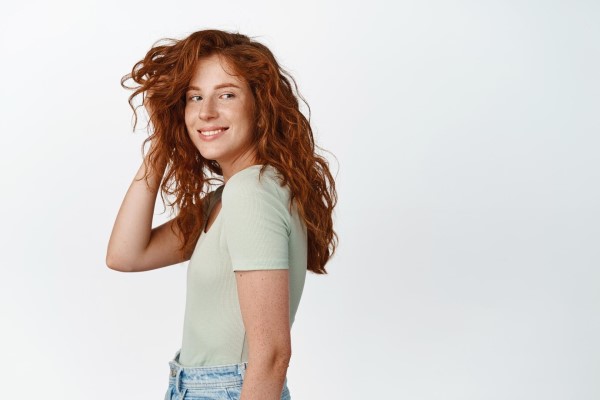 Linda chica con melena rizada roja y pecas jugando con corte de pelo y sonriendo feliz a la cámara de pie en camiseta