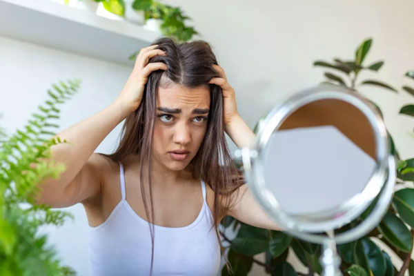 Tricofagia - Joven de ojos claros se observa preocupada frente al espejo por pérdida sostenida de cabello en el área frontal de la cabeza 