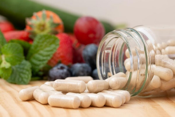 Frasco de vidrio con cápsulas de vitaminas caen sobre una superficie de madera con fresas y arándanos de fondo