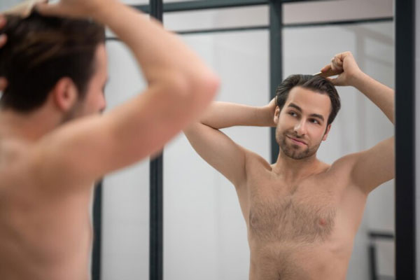 Shock loss tras injerto capilar - Joven atractivo y sin camiseta contempla su imagen mientras peina su cabello frente al espejo
