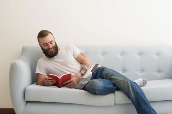 Hombre con barba lee un libro mientras descansa un rato sobre el sofá