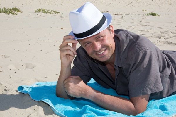 Hombre acostado en una toalla sobre la arena de la playa tocando su sombrero con la mano