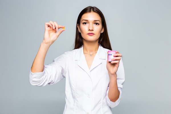 Mujer joven de pelo negro con bata blanca muestra un recipiente para pastillas y una pastilla en su mano