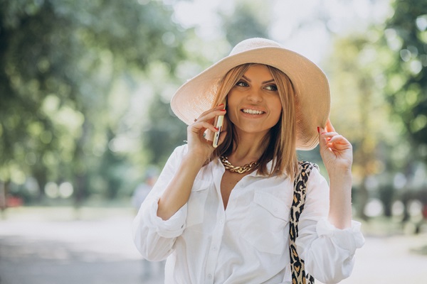 Síndrome de ovario poliquístico - Mujer joven usando un teléfono mientras toca su sombrero en un parque