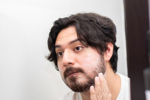 Joven de abundante cabello se mira al espejo mientras se aplica crema de afeitar en la barba
