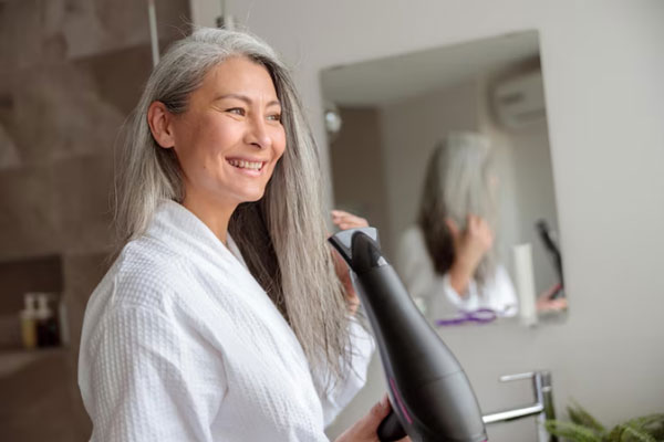 Mujer canosa en bata de baño utiliza el secador para retirar la humedad de su cabello