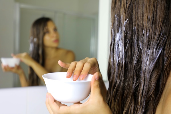 Mujer frente al espejo sostiene en sus manos un recipiente mientras aplica una mascarilla capilar
