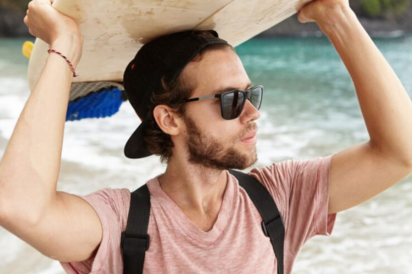 Joven con lentes oscuros utiliza una gorra y una tabla del surf para protegerse de los rayos del sol