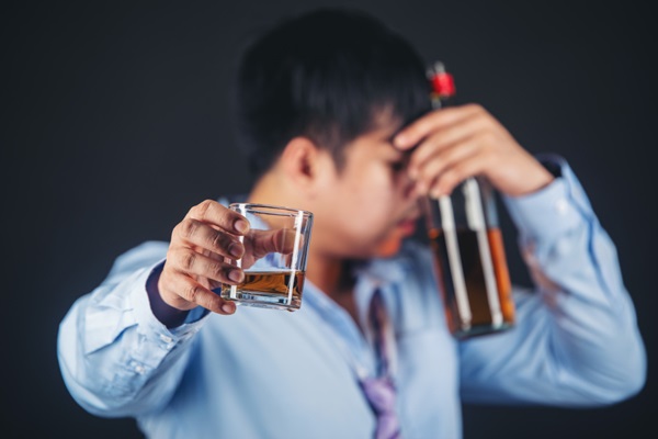Hombre alcohólico tapándose la cara con una botella mientras sostiene en su mano un vaso de whisky