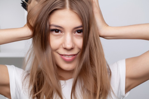 Mujer joven de cabello castaño, liso y brillante sonriendo con las manos en la cabeza