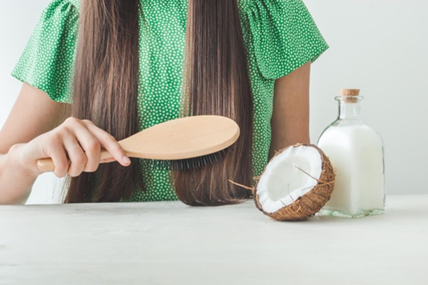 Cómo cuidar el cabello mojado - Mujer peinando su cabello con un cepillo de madera junto a un recipiente de aceite de coco que ayuda a hidratar el pelo