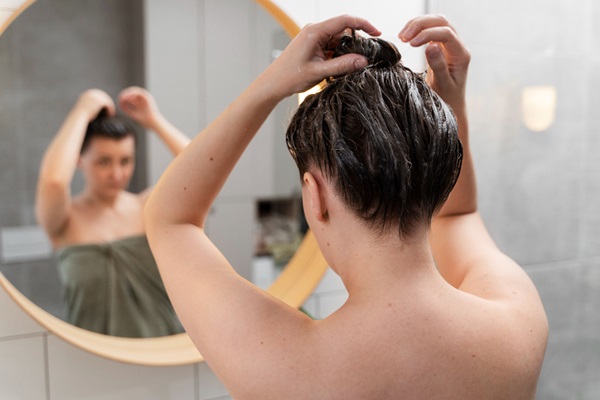 Cómo cuidar el cabello mojado - Mujer observándose en el espejo al recoger su cabello húmedo después de lavarlo, aumentando el riesgo de quebrar las hebras capilares