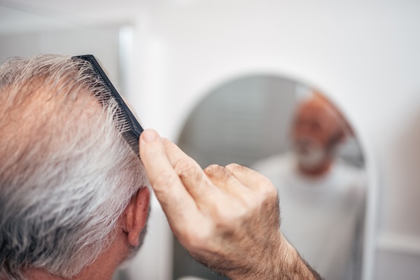 Injerto de pelo con canas - Hombre con pelo canoso observándose en el espejo mientras peina su cabello con alopecia en la coronilla