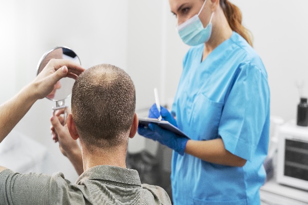 etapas del implante capilar, hombre con evidente alopecia sistene un espejo mientras se toca la linea del cabello, todo esto frente a especialista que anota sus observaciones 